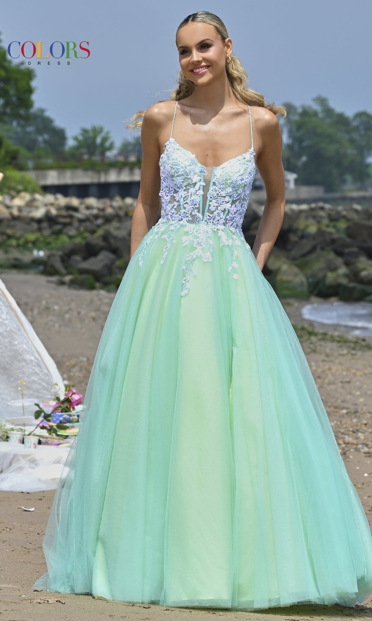 A woman stands on a beach wearing the Colors Dress 3497, a strappy-back prom ball gown in mint green with a white sheer floral lace bodice and delicate thin straps.